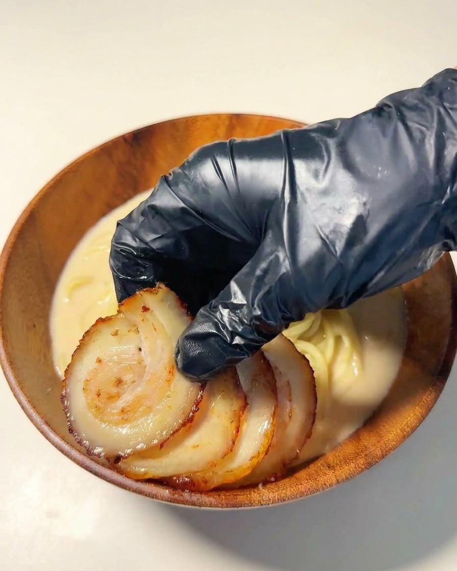Eine Hand mit schwarzem Handschuh legt Scheiben von gebratenem Chashu auf die Ramen-Nudeln.