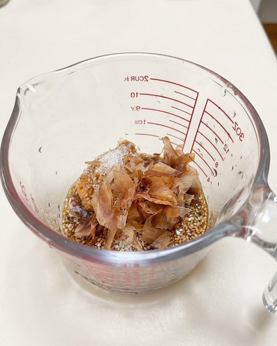 Bonitoflocken liegen auf der Misopaste und dem Sesam in einem Glasmessbecher.