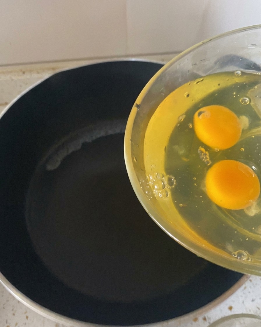 Zwei rohe Eier werden aus einer Glasschüssel in eine heiße, dunkle Bratpfanne gegossen.