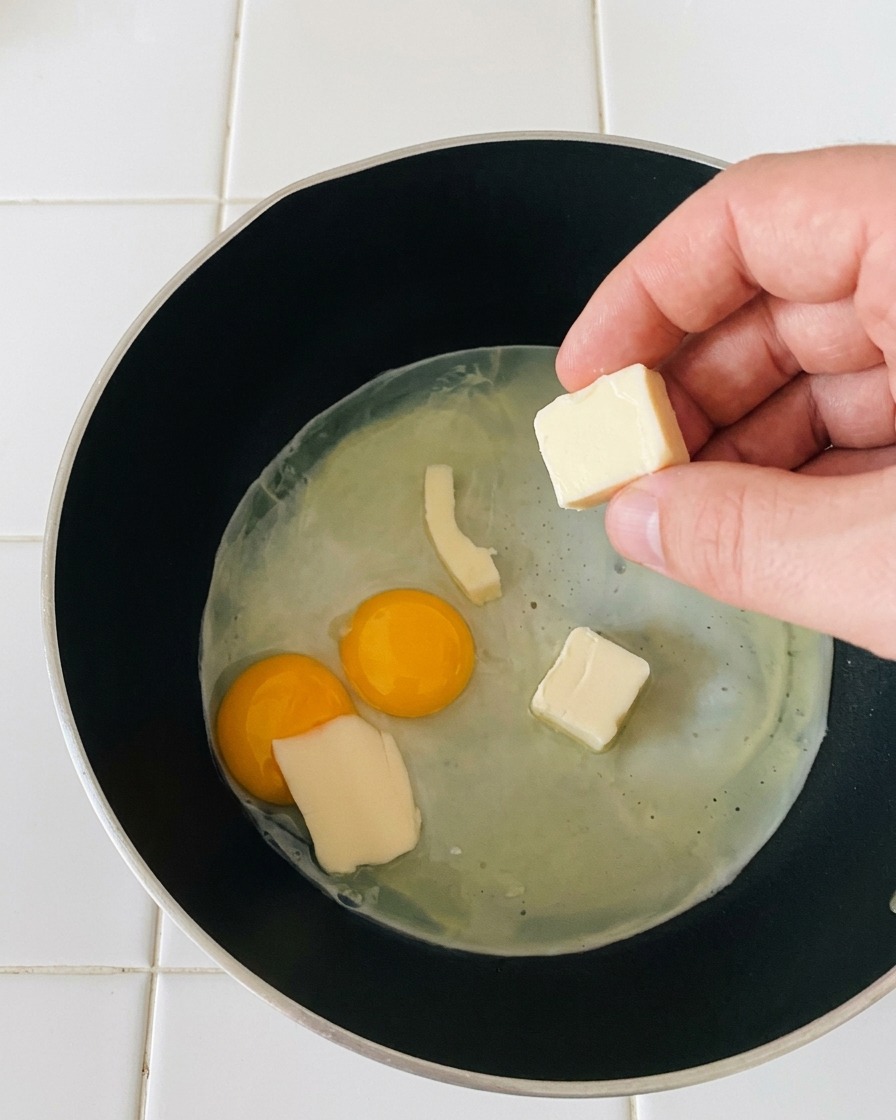 Eine Hand legt kleine, rechteckige Scheiben weißen Mozzarellas auf die sanft bratenden Spiegeleier.