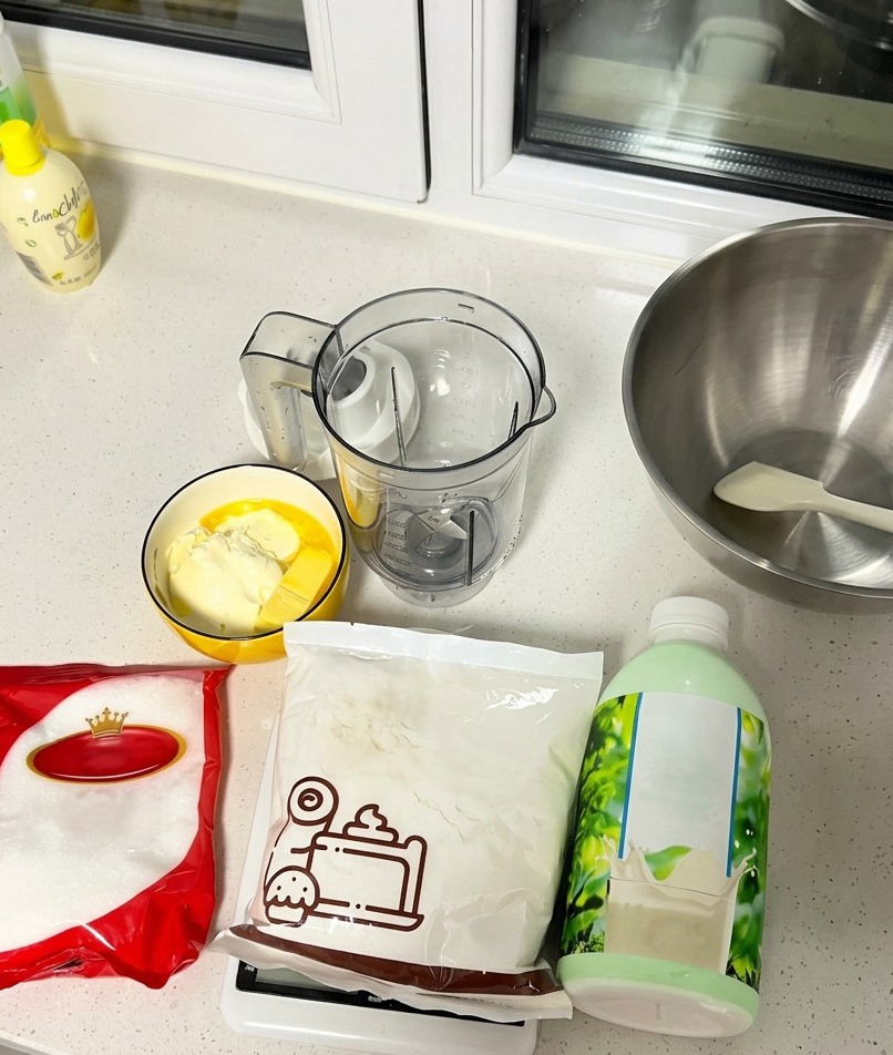 Eine Auswahl an Backzutaten wie Milch, Zucker, Mehl, Butter und Frischkäse auf einer Arbeitsfläche.