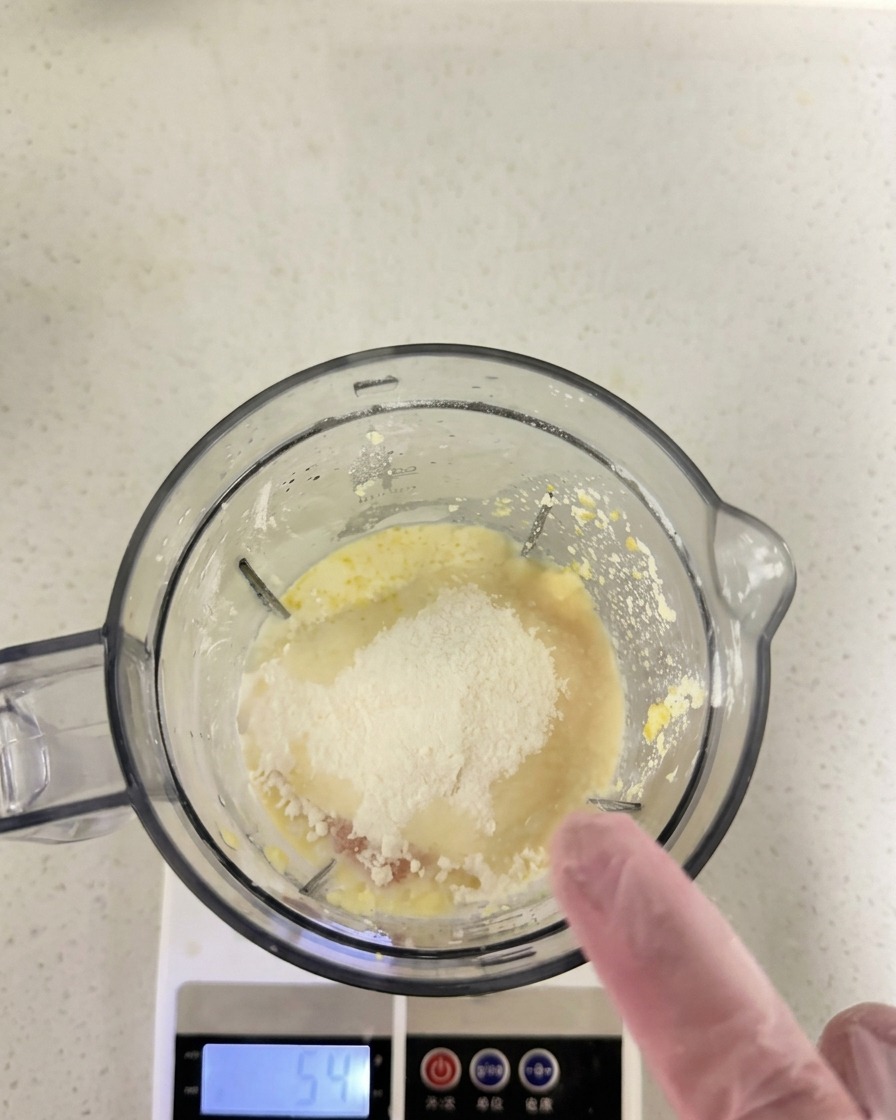 Mehl und trockene Zutaten auf der flüssigen Mischung in einem Mixerbecher, der auf einer Digitalwaage steht.