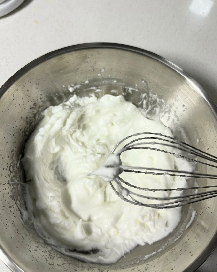 Ein Schneebesen hebt fluffigen Eischnee aus einer Schüssel, der weiche Spitzen bildet.