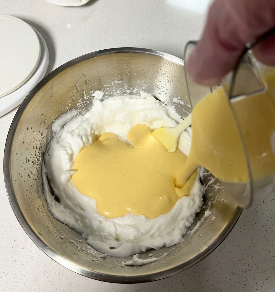 Glatte gelbe Käsemasse wird in eine Schüssel mit fluffigem weißem Eischnee gegossen.