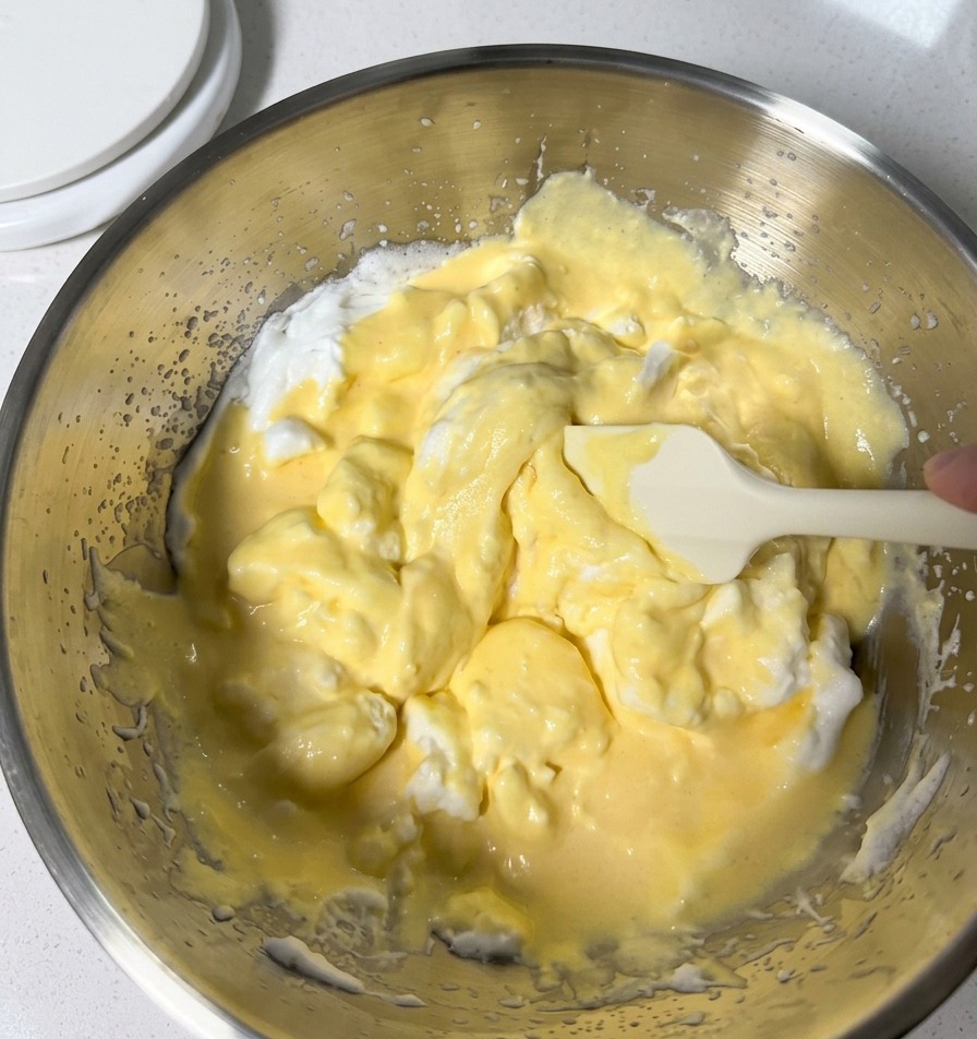 Vorsichtiges Unterheben von hellgelbem Teig und weißem Eischnee mit einem Silikonspatel in einer Metallschüssel.
