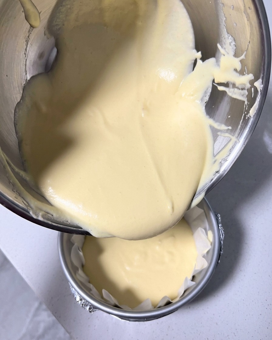Hellgelber Käsekuchenteig wird aus einer Schüssel in eine runde, mit Backpapier ausgekleidete Form gegossen.