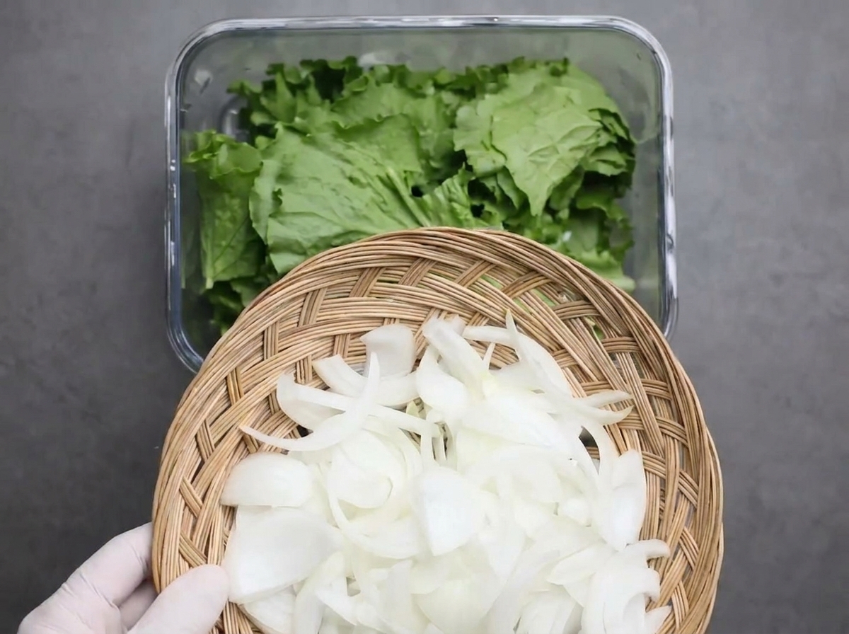 Geschnittene weiße Zwiebeln werden über den gezupften Kopfsalat in einen Glasbehälter gestreut.