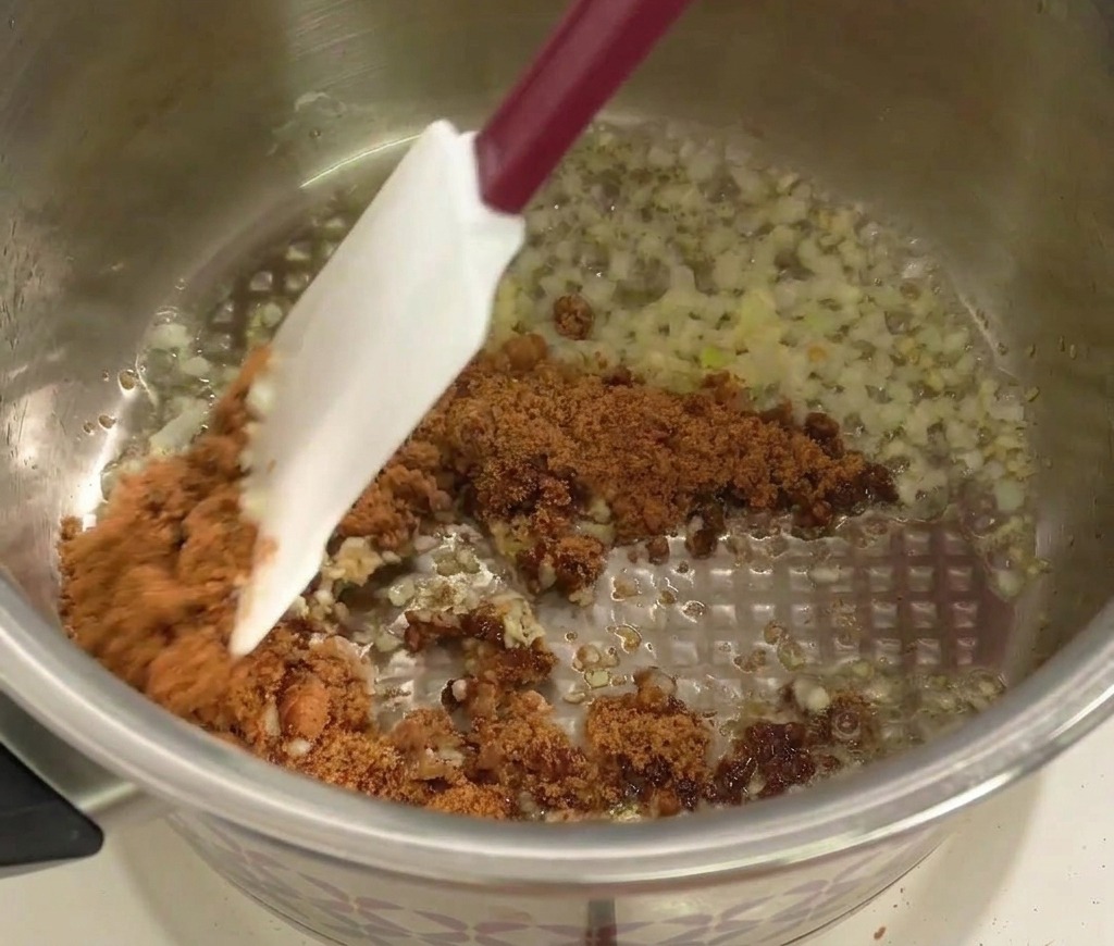 Ein Spatel rührt braunen Zucker unter die angedünsteten Zwiebeln und den Knoblauch im Topf.