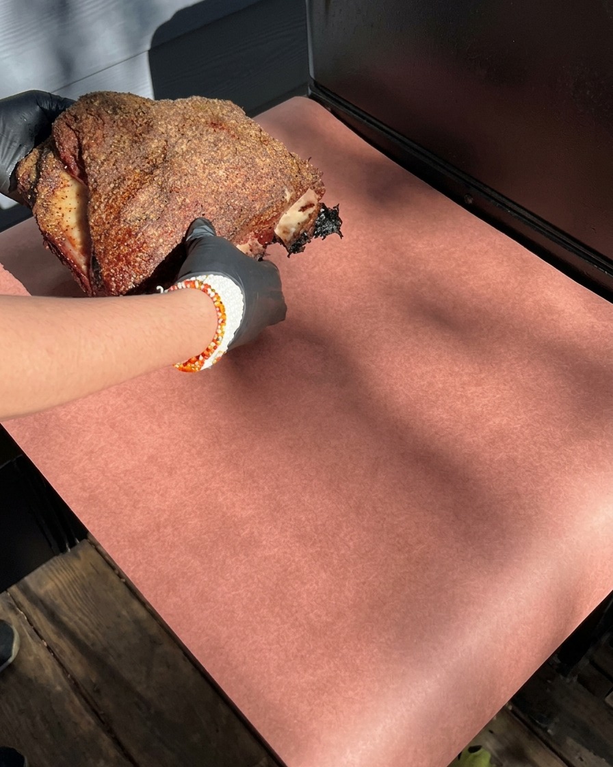 Hände in Handschuhen halten eine große geräucherte Rinderrippe mit dunkler Gewürzkruste und legen sie auf rosa Metzgerpapier.