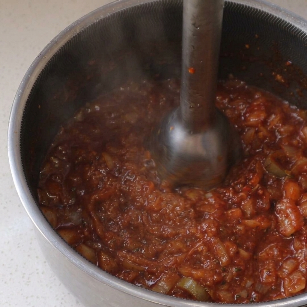 Mit einem Pürierstab wird eine köchelnde, stückige rote BBQ-Sauce in einem Edelstahltopf fein püriert.