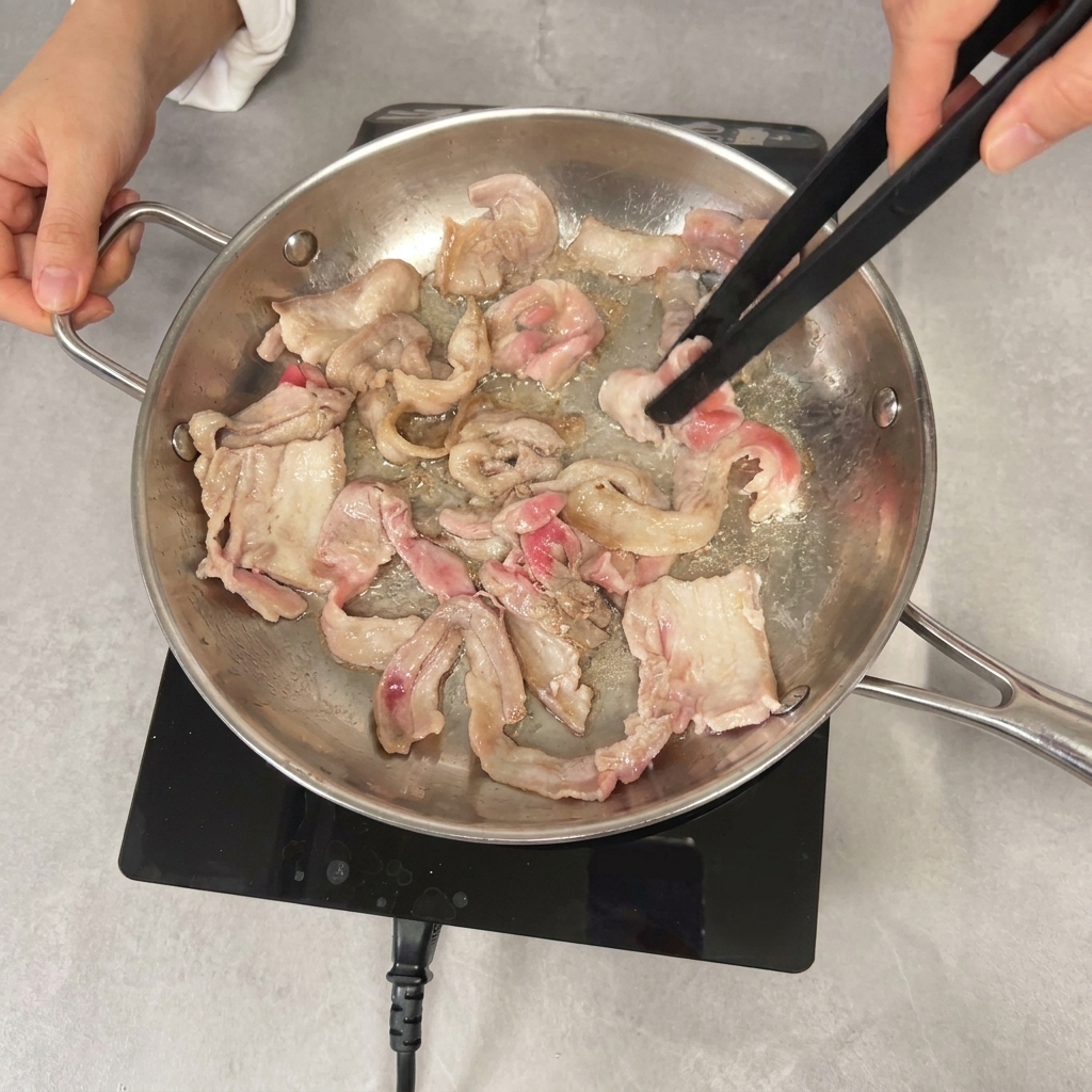 Eine Grillzange wendet leicht gebräunte Schweinebauchscheiben in der Pfanne.