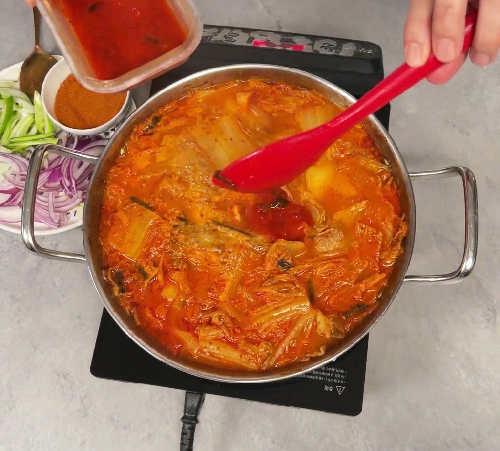 Roter Kimchi-Saft wird aus einem kleinen Behälter in den köchelnden Eintopf gegossen.