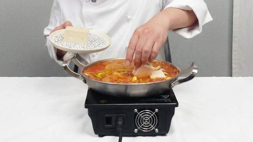 Weiße Tofuscheiben werden vorsichtig in den roten Kimchi-Eintopf gelegt.