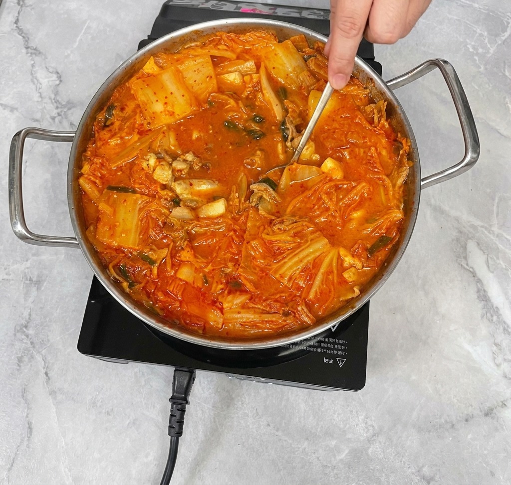 Ein Edelstahllöffel rührt vorsichtig im leuchtend roten Kimchi-Eintopf.