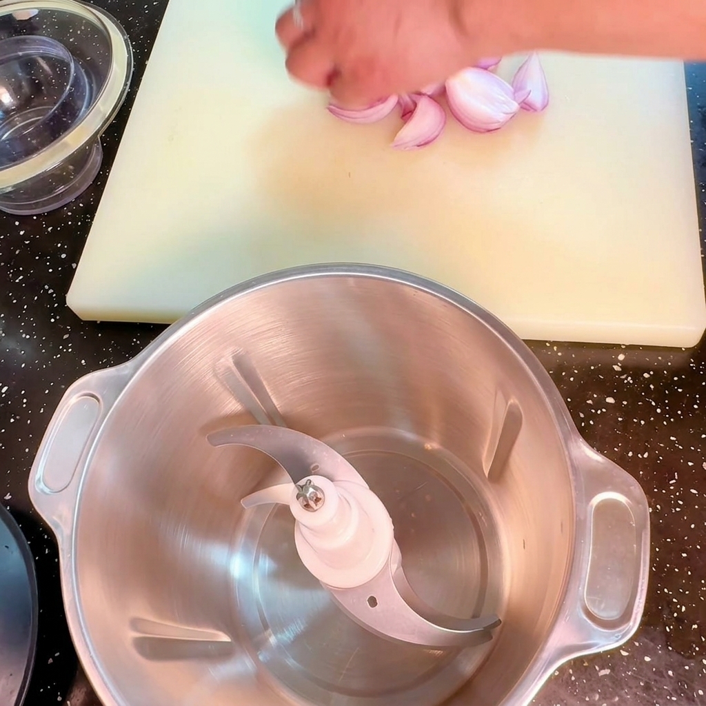 Eine Hand legt gehackte rote Zwiebeln neben einen Edelstahl-Mixbehälter auf einem weißen Schneidebrett.