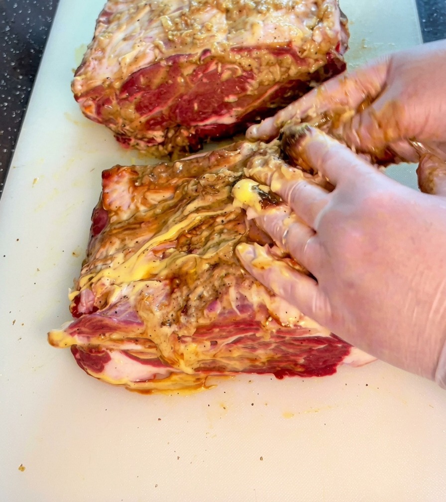 Hände massieren eine Mischung aus Saucen und Gewürzen in ein dickes Stück rohes Rindfleisch auf einem weißen Brett.