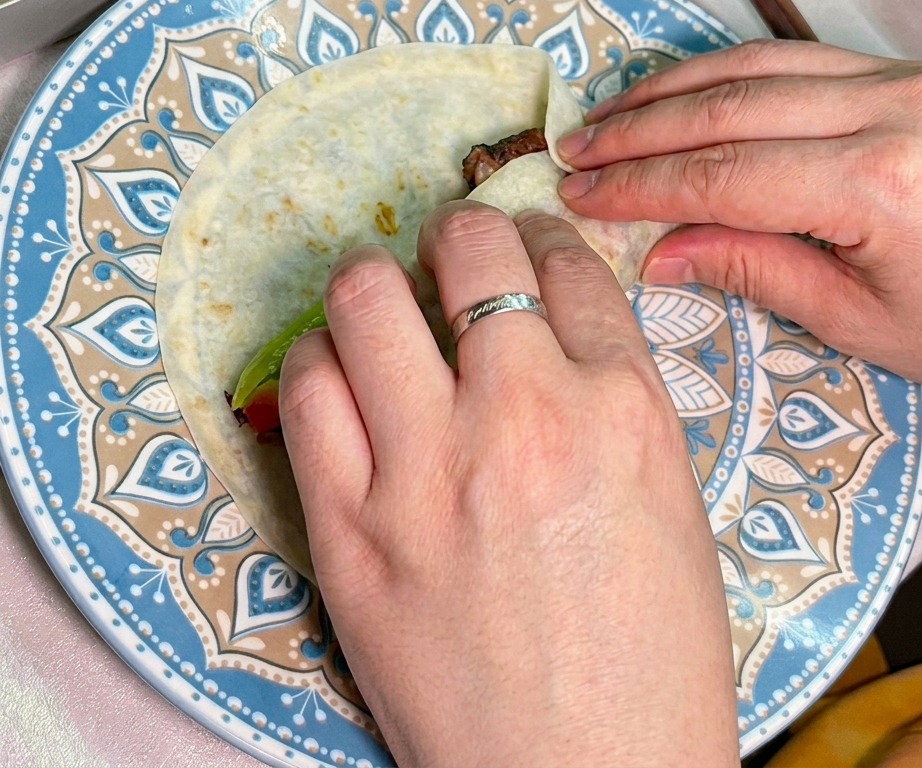 Zwei Hände rollen eine mit gesmoktem Rindfleisch und frischem Gemüse gefüllte Tortilla sorgfältig zusammen.