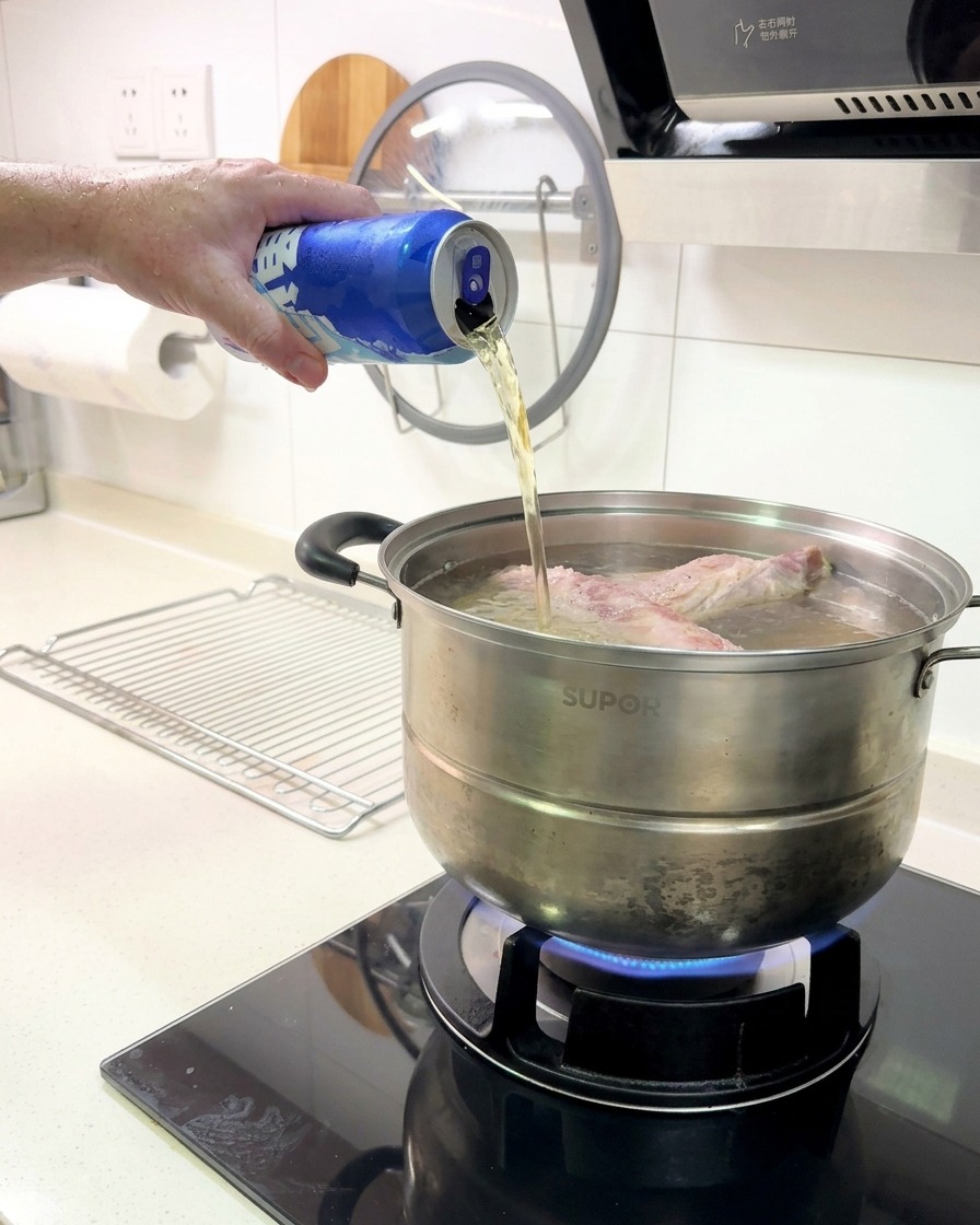 Bier wird aus einer blauen Aluminiumdose in einen Topf mit Rippchen gegossen, die auf einem Gasherd kochen.
