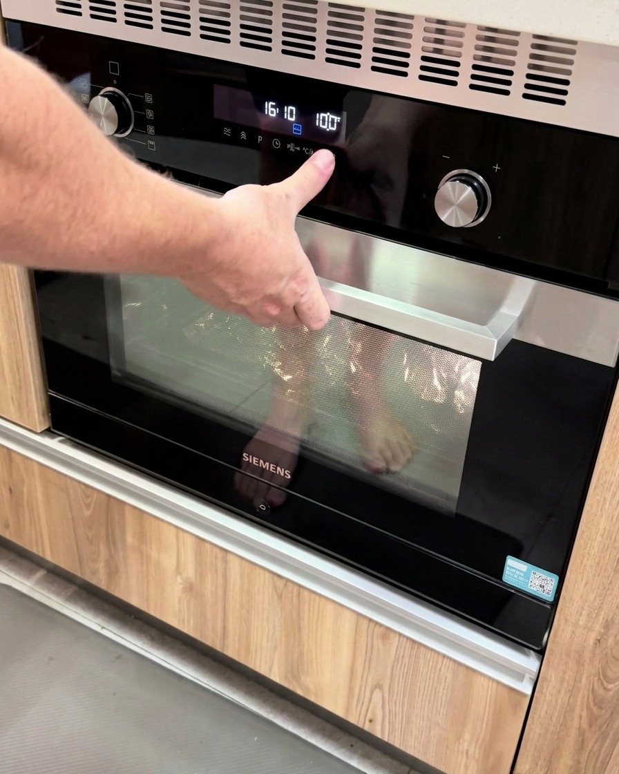 Eine Hand drückt eine Taste auf einem modernen digitalen Ofenfeld, auf dem die Temperatur auf 100 Grad eingestellt ist.