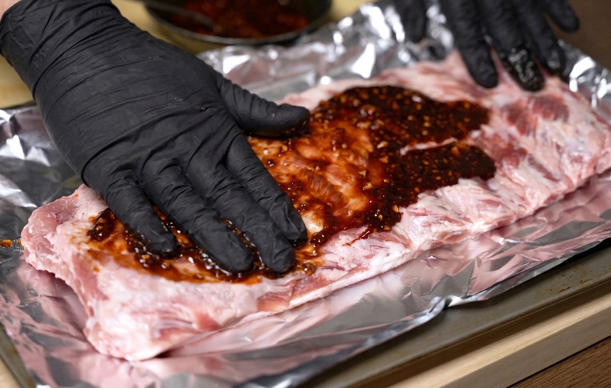 Hände in Handschuhen reiben eine dicke rote Marinade gleichmäßig auf die rohen Rippchen auf Alufolie.