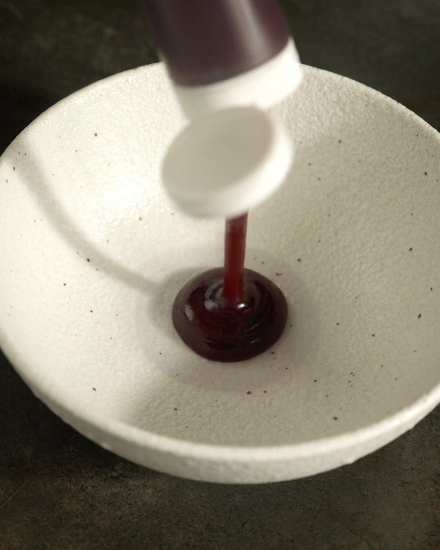 Dunkle BBQ-Sauce wird aus einer Flasche in eine weiße Keramikschale gegeben.