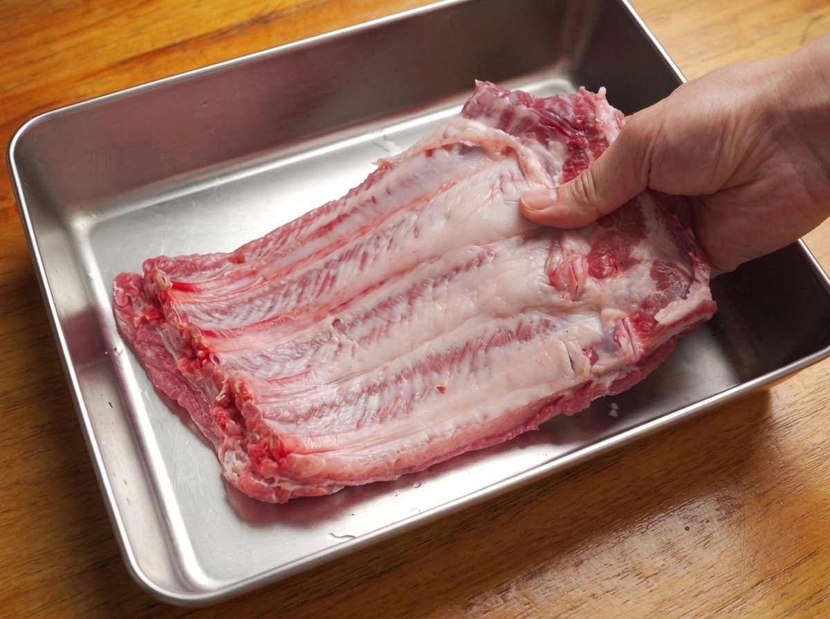 Hände ziehen die Silberhaut von der Rückseite einer großen Schweinerippchen-Seite in einer Metallschale ab.