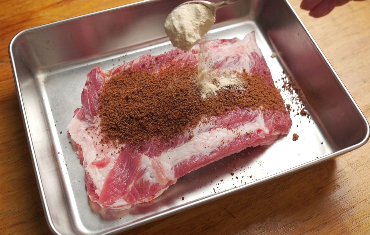 Rohe Schweinerippchen werden mit braunem Zucker und einer Gewürzmischung von einem Löffel bestreut.