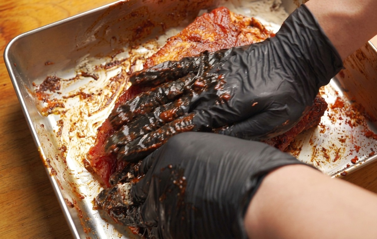 Hände mit Handschuhen massieren eine dicke, rötlich-braune Marinade in die Schweinerippchen ein.