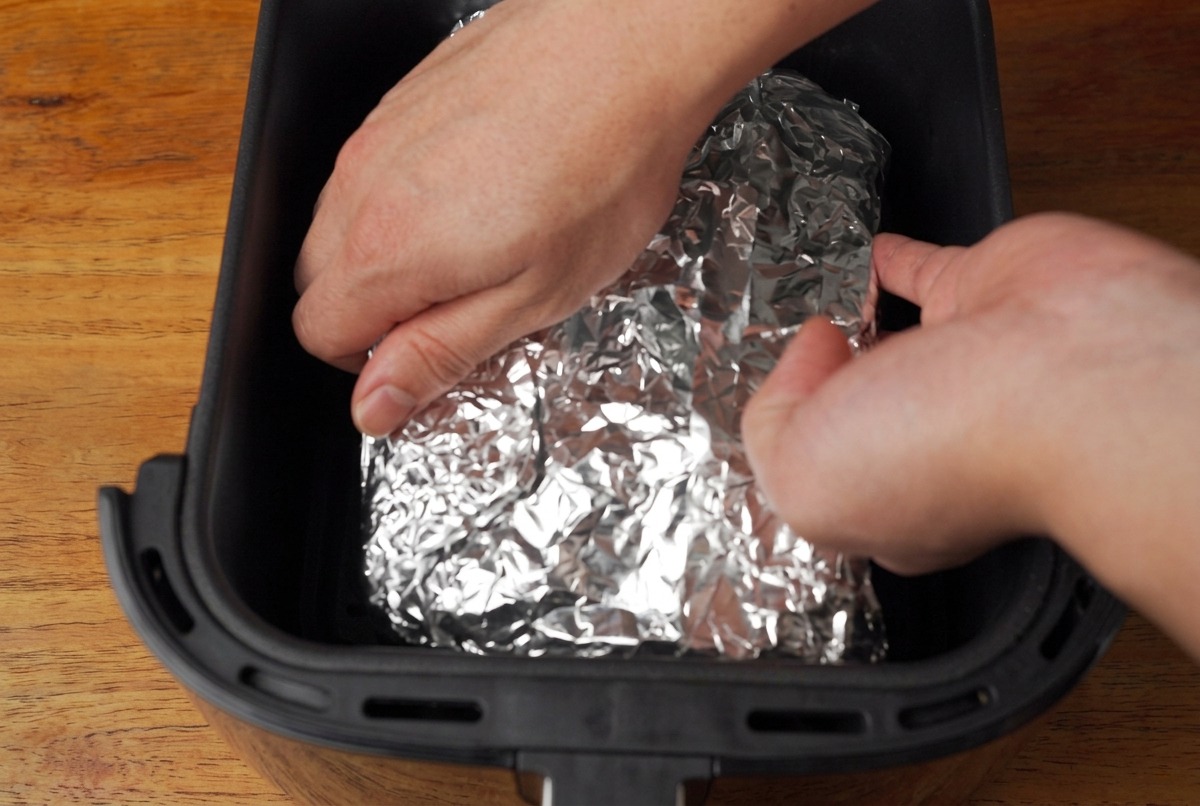 Hände legen das in Folie gewickelte Paket in den schwarzen Korb einer Heißluftfritteuse.