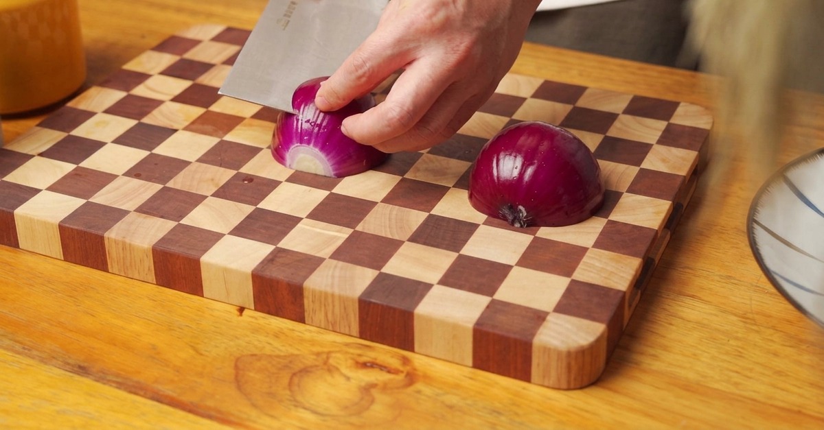 Hände schneiden eine rote Zwiebel auf einem Holzbrett in feine Würfel.