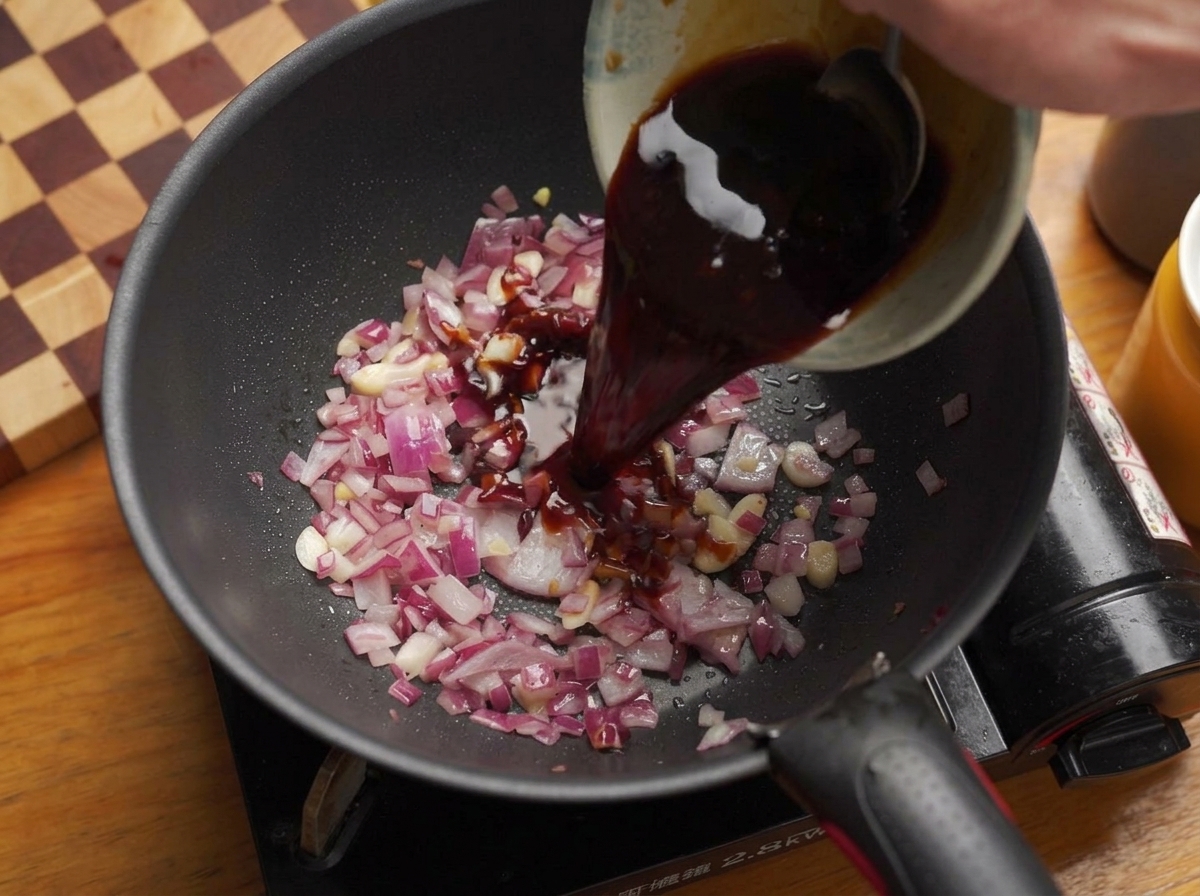 Eine dunkle Saucenmischung wird aus einer Schüssel in die Pfanne zu den Zwiebeln gegossen.