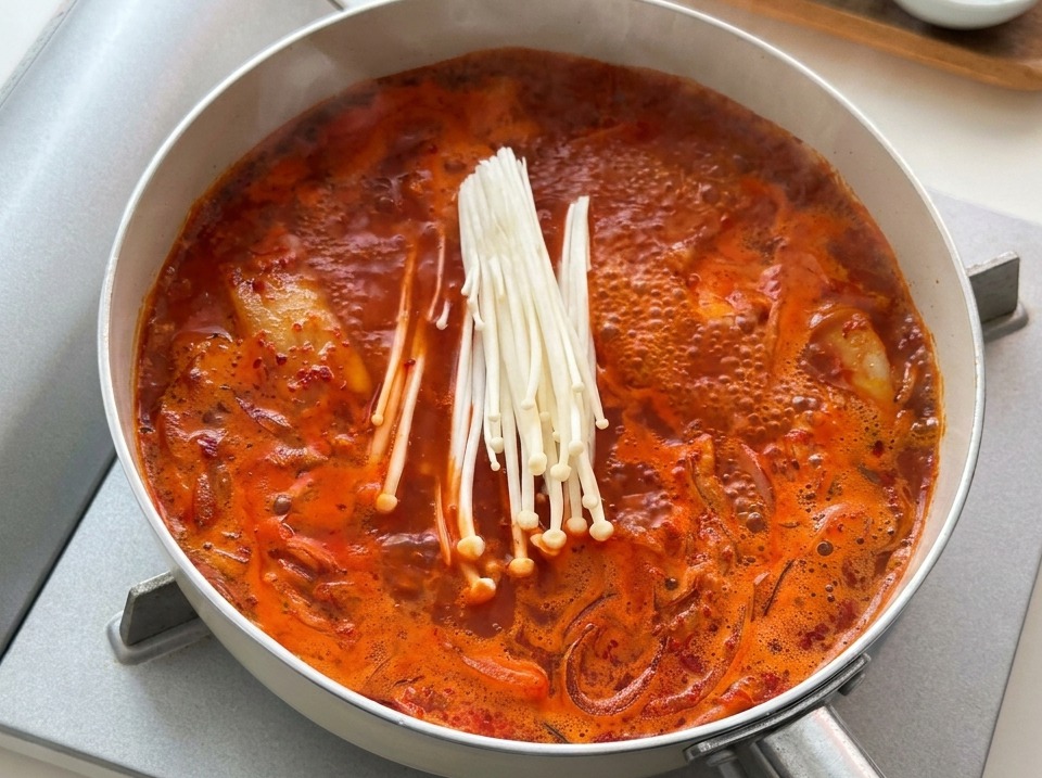 Ein Bund weißer Enoki-Pilze wird in die kochende rote Kimchi-Suppe gelegt.