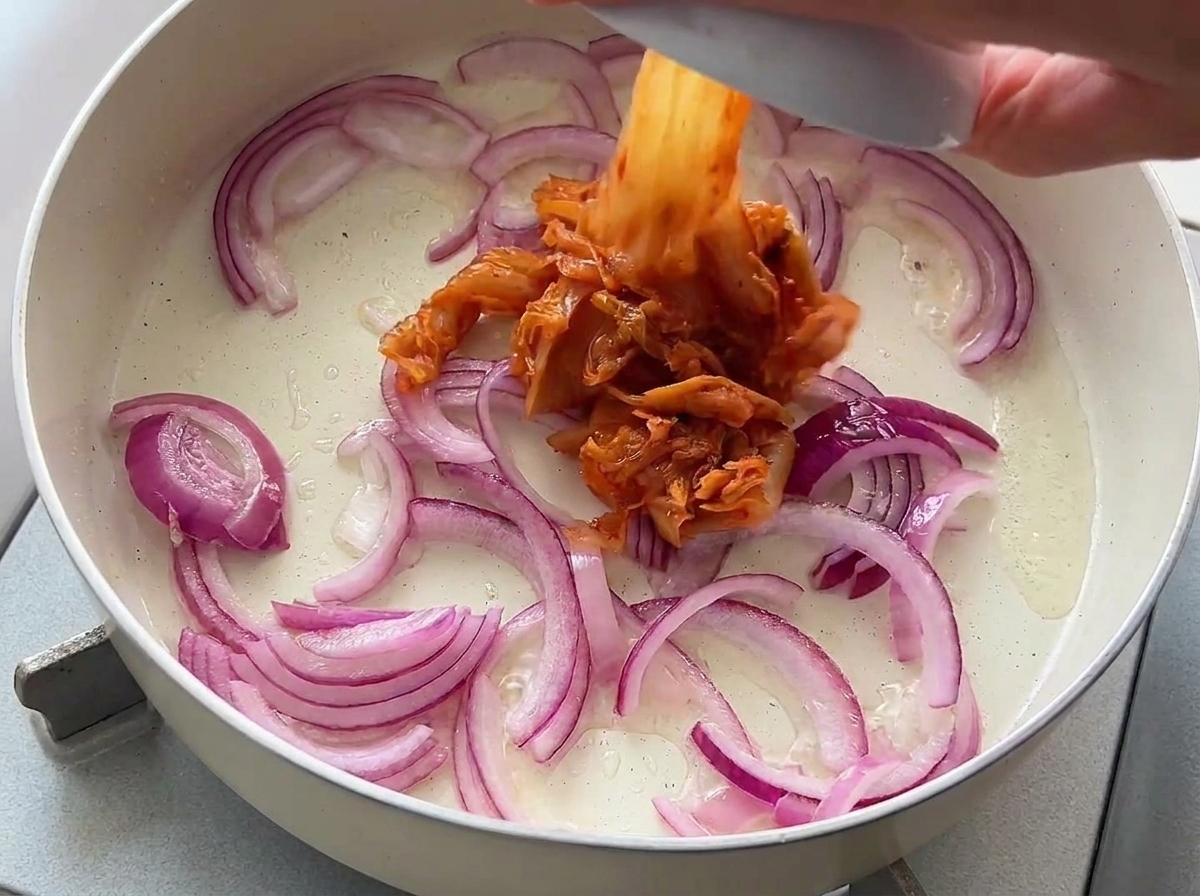 Gehacktes Kimchi wird aus einer kleinen Schüssel zu den angebratenen Zwiebeln in die Pfanne gegeben.