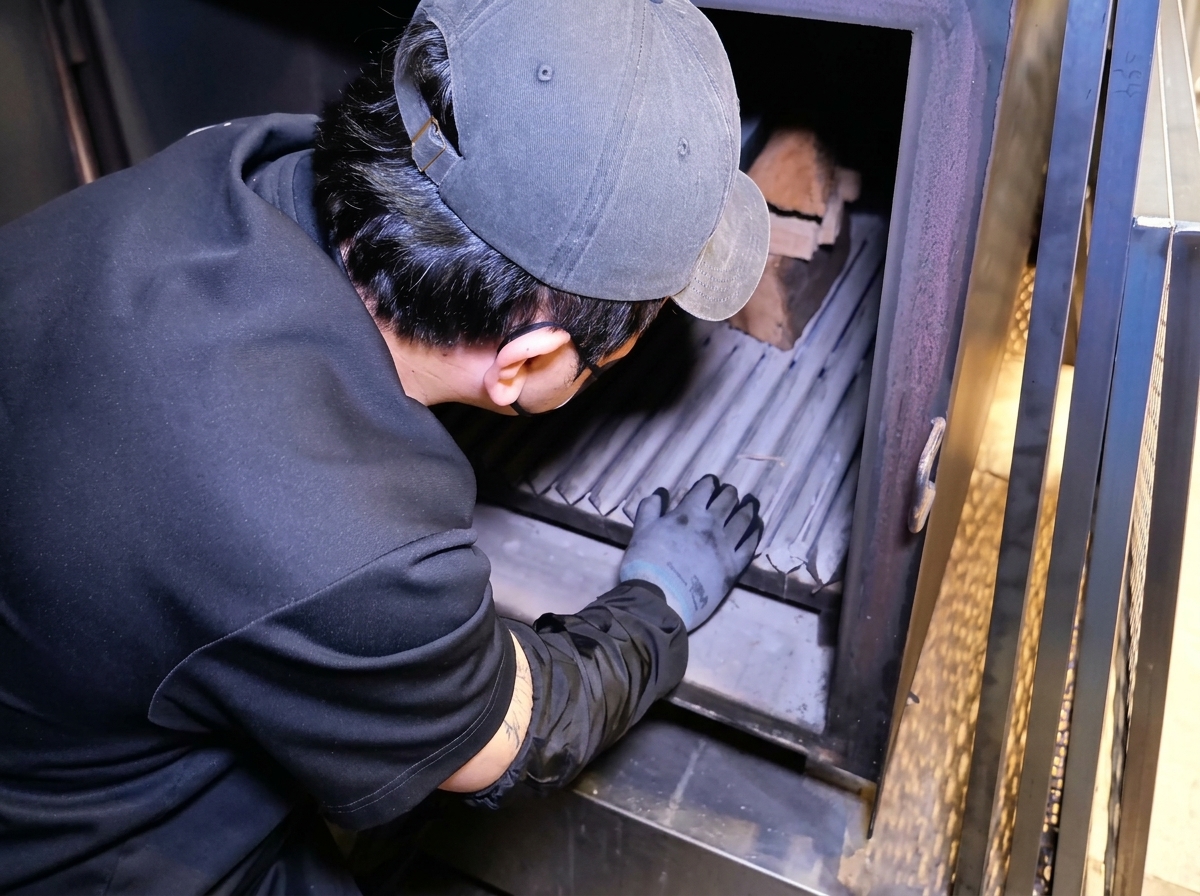 Eine Person in schwarzer Kleidung arrangiert Holzscheite in der Feuerbox eines großen Metall-Smokers.