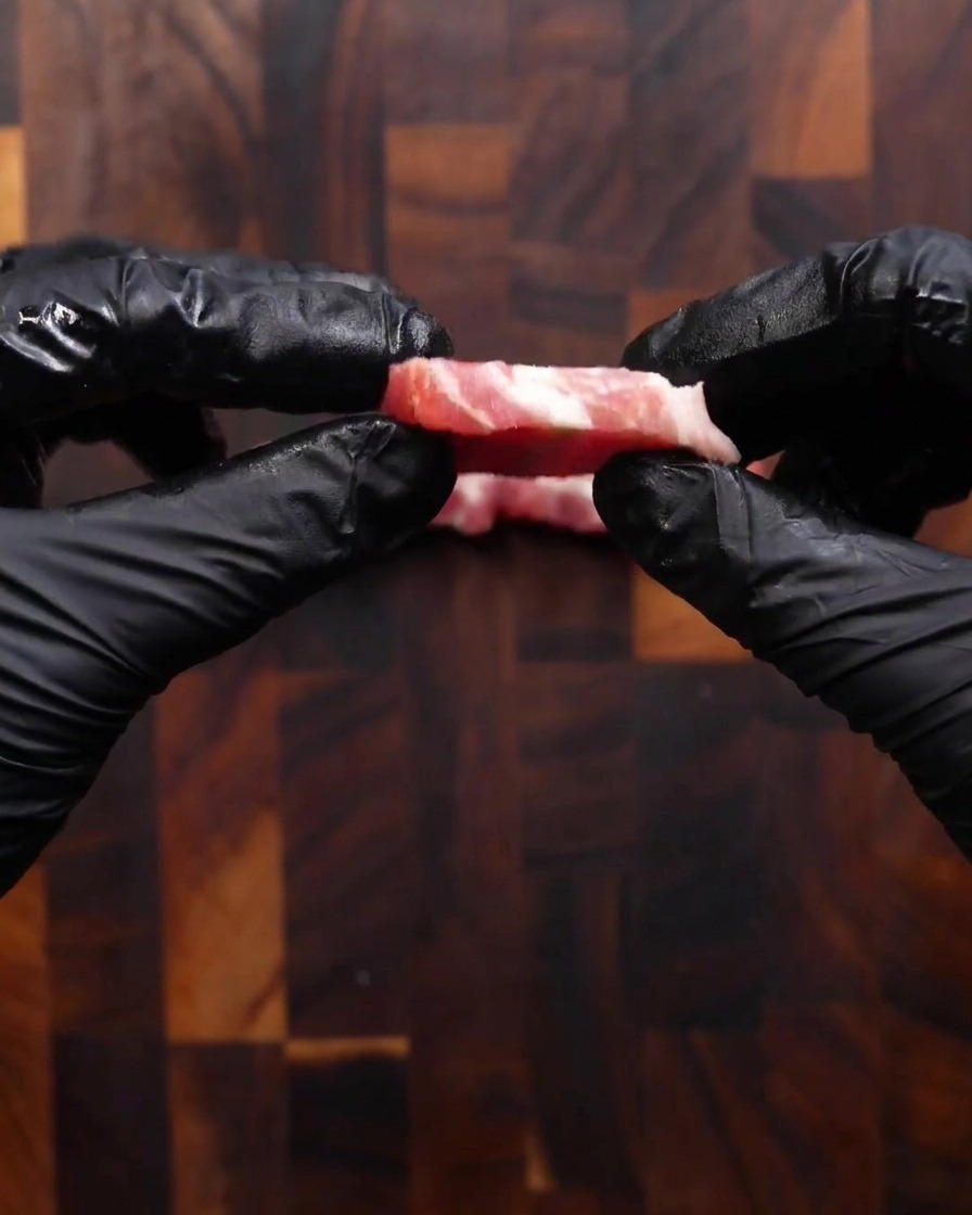 Zwei Hände in schwarzen Handschuhen zeigen eine frisch geschnittene Scheibe rohen Schweinebauch, die etwa so dick wie eine Münze ist.