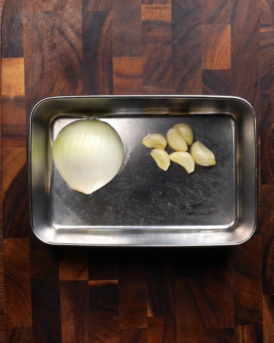 Ein Edelstahltablett mit einer halben weißen Zwiebel und mehreren geschälten Knoblauchzehen auf einem Holzbrett.