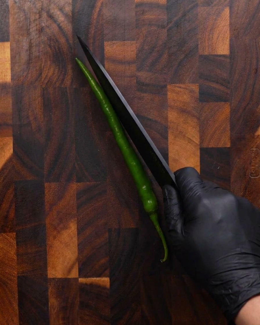 Eine Hand schneidet eine lange grüne Chilischote diagonal mit einem scharfen schwarzen Messer auf einem Holzbrett.