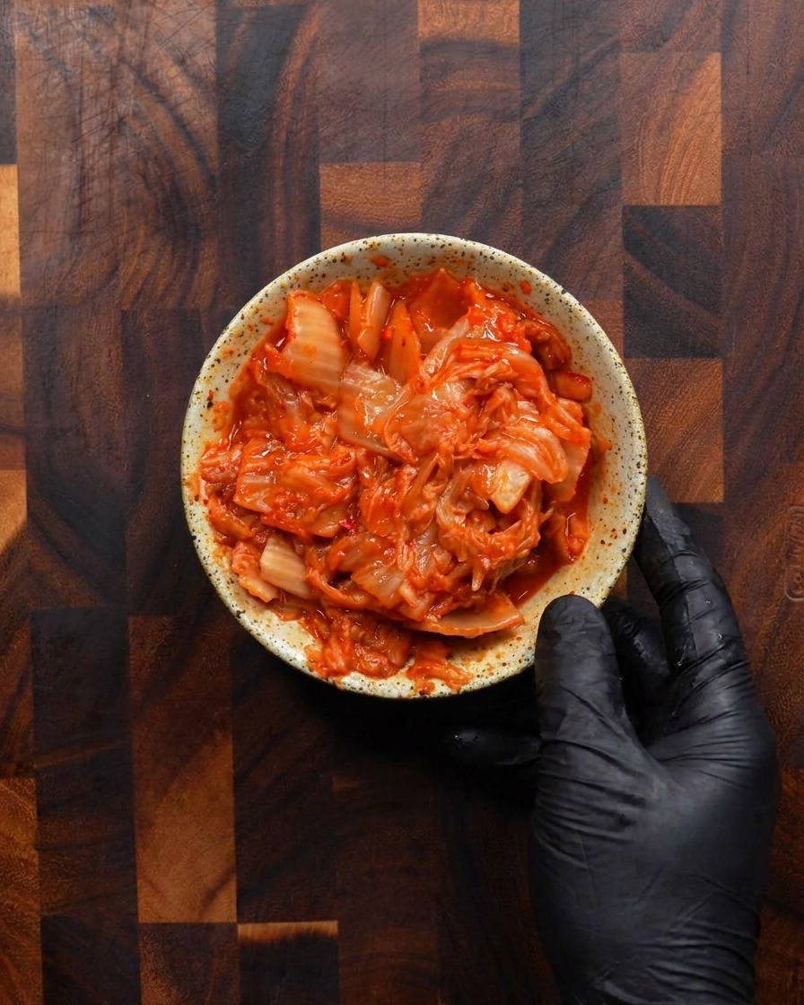 Eine Hand hält eine rustikale Schale voll mit leuchtend rotem, grob gehacktem fermentiertem Kimchi.