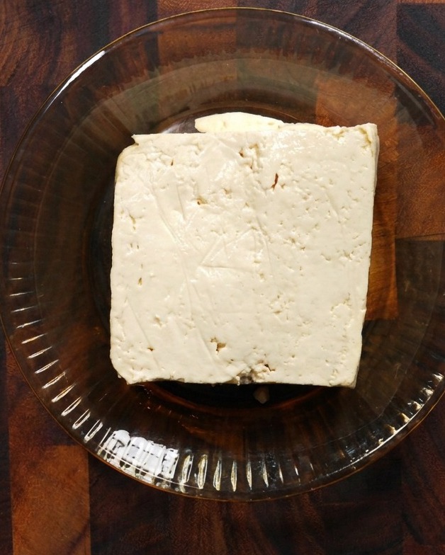 Ein quadratischer Block fester weißer Tofu liegt in einer flachen Glasschale auf einer Holzoberfläche.