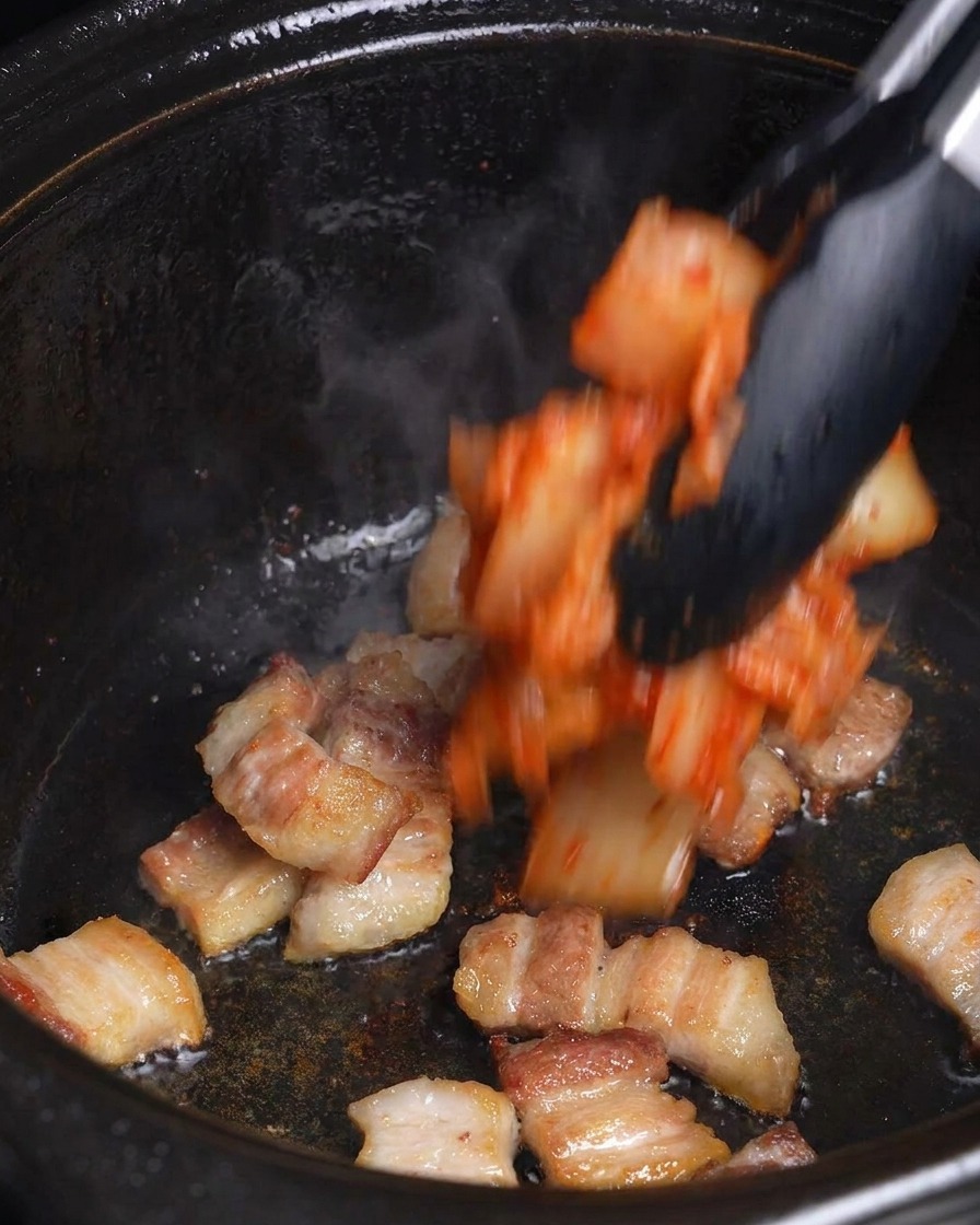 Gehacktes rotes Kimchi wird mit einer Zange zu dem gebräunten Schweinebauch in einen schwarzen Topf gegeben.