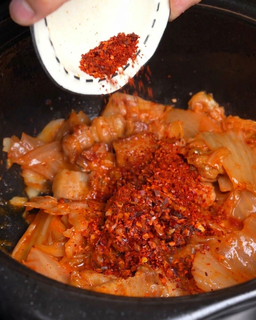 Rotes Chilipulver wird über das angebratene Kimchi und den Schweinebauch in einen schwarzen Topf gestreut.