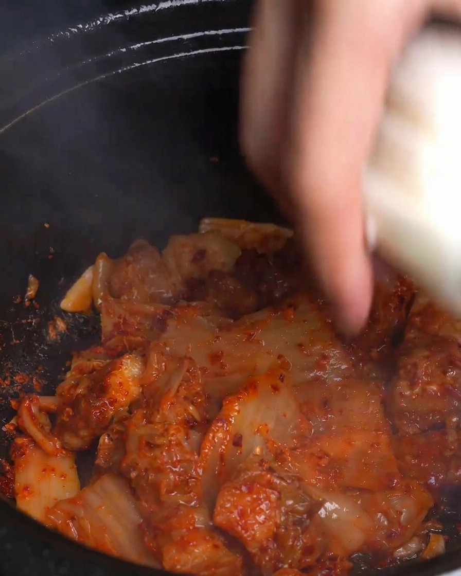 Eine Hand gibt dicke weiße Zwiebelscheiben in den dampfenden Topf mit rotem Kimchi und Schweinebauch.