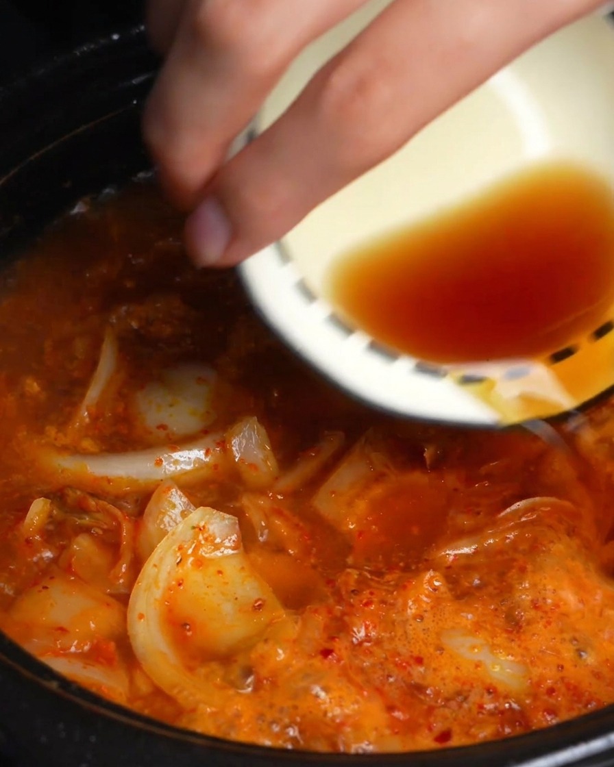 Eine Hand gießt Soja- und Fischsauce aus einer Schale in die köchelnde rote Kimchi-Brühe.
