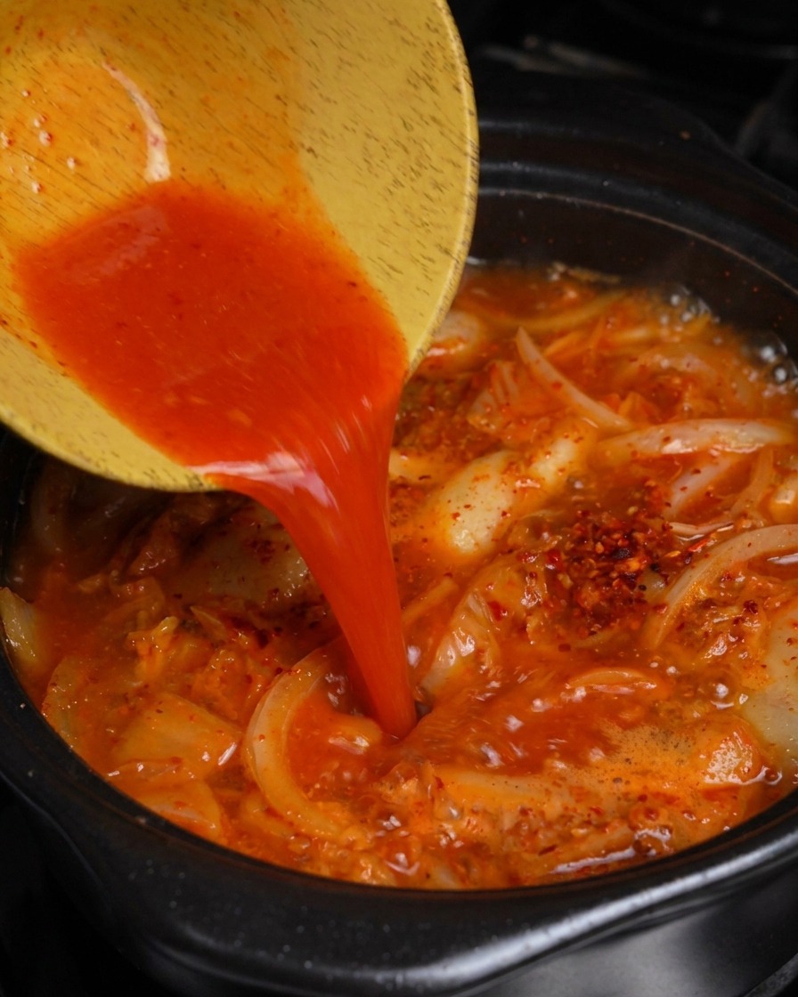 Leuchtend roter Kimchi-Saft wird aus einer Holzschale in den sprudelnden Eintopf gegossen.