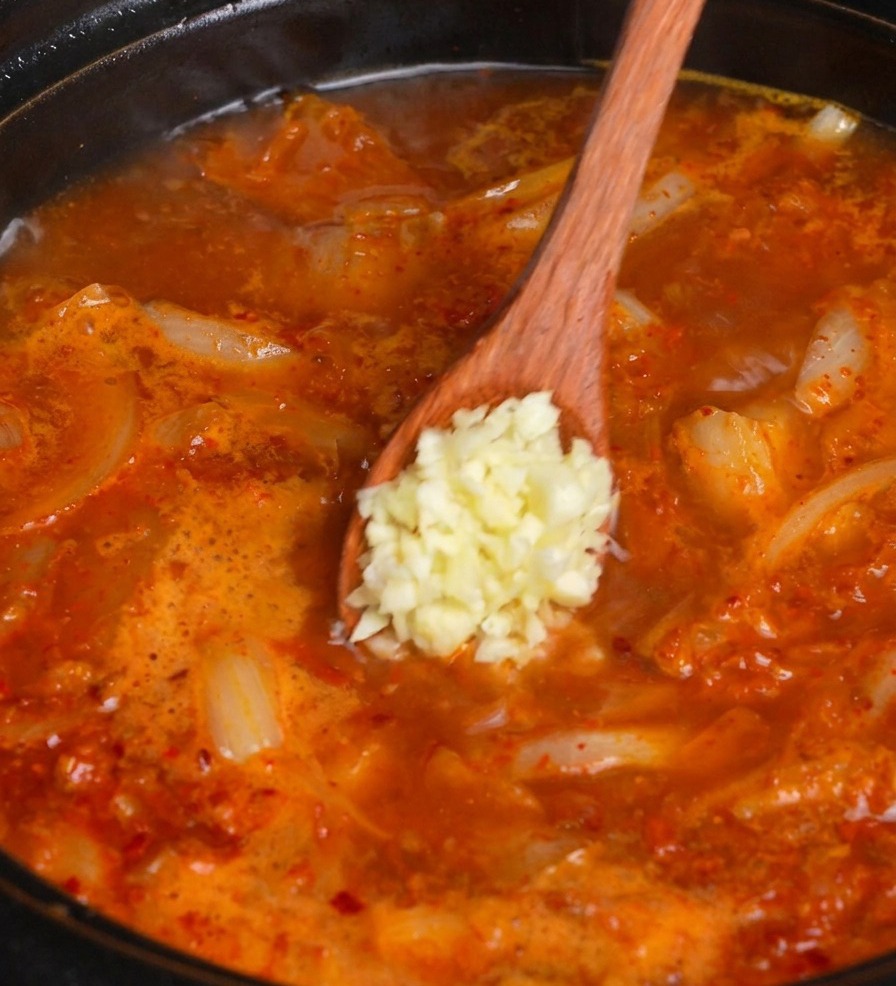 Ein Holzlöffel gibt gehackten Knoblauch in den köchelnden Topf mit roter Kimchi-Suppe.