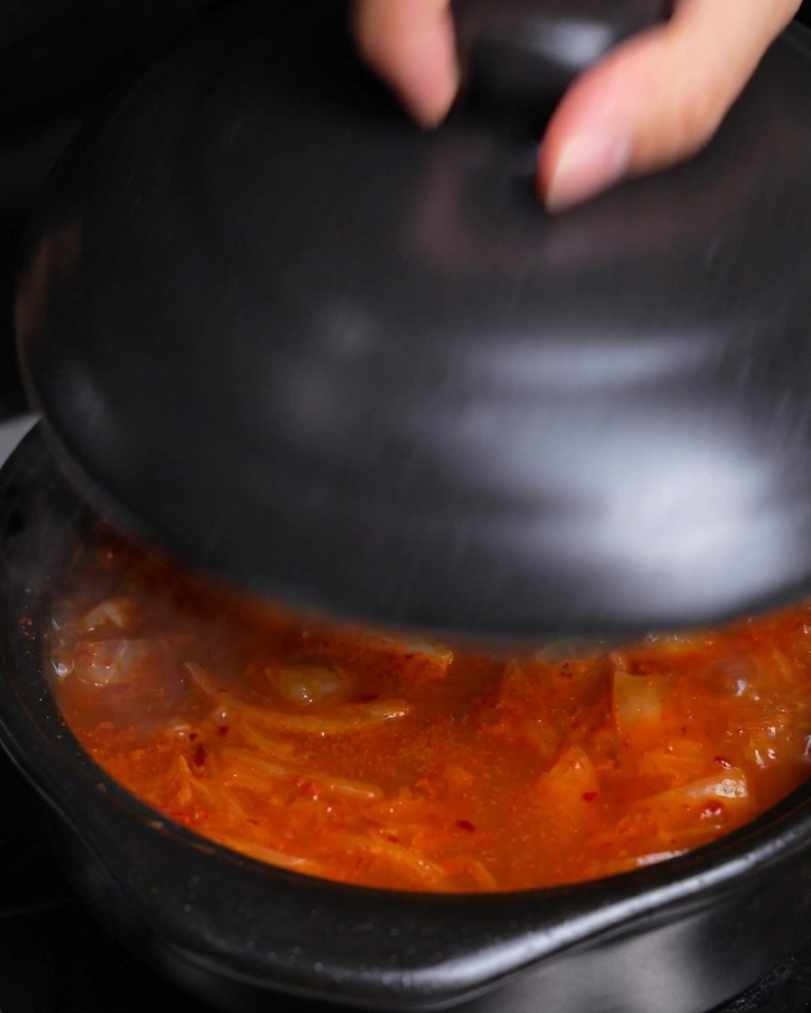 Eine Hand setzt einen Keramikdeckel auf den Topf mit kochendem Kimchi-Eintopf.