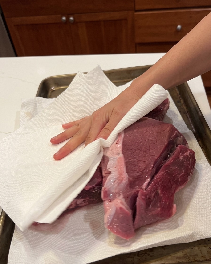 Eine Hand tupft ein großes Stück rohes Rindfleisch auf einem Backblech mit einem Papiertuch trocken.