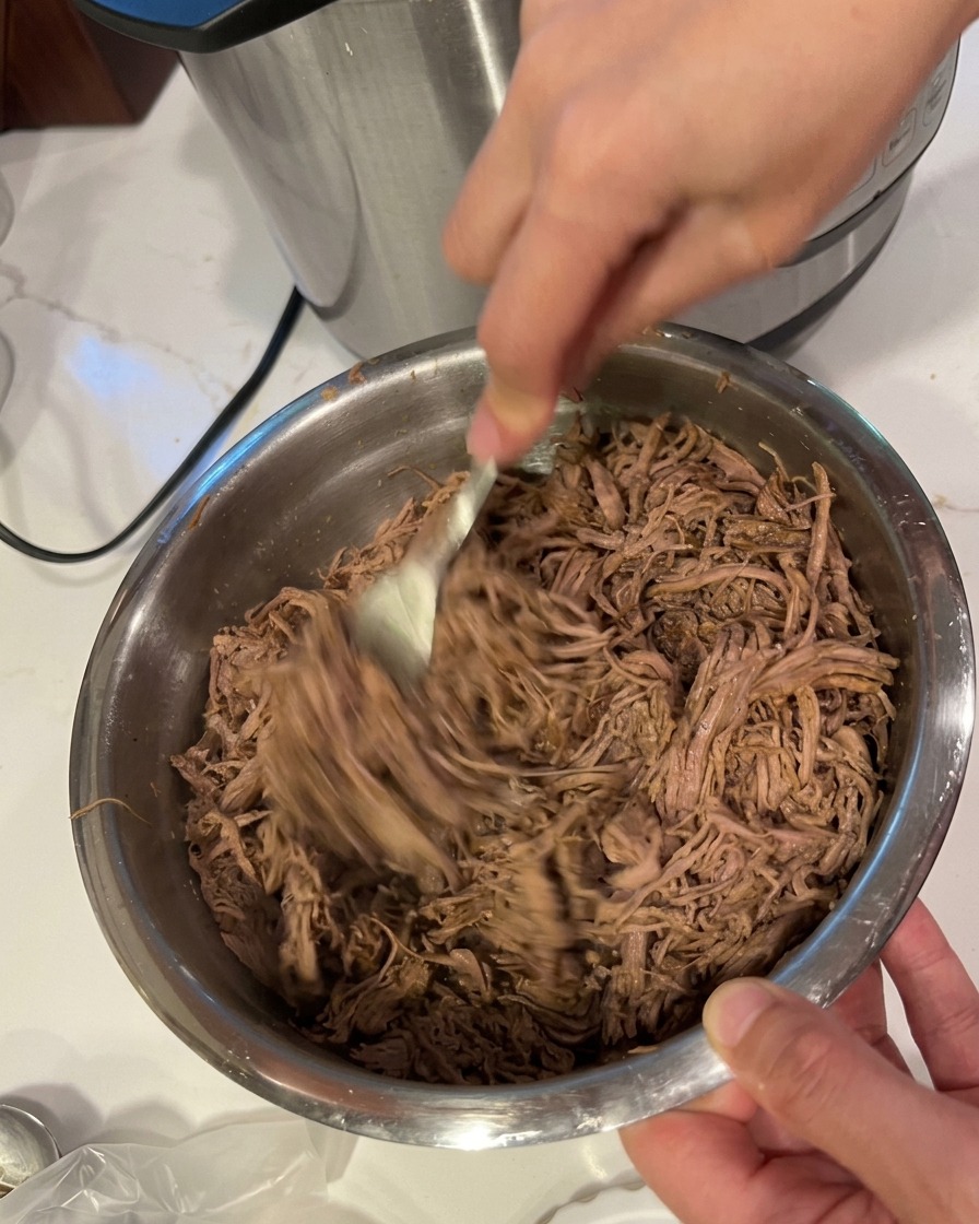 Hände zerzupfen das zarte Rindfleisch mit zwei Gabeln in feine Fasern in einer Edelstahlschüssel.