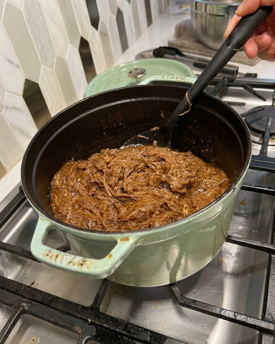 Gezupftes Rindfleisch wird mit einem Teigschaber unter die dunkle BBQ-Sauce im Topf gehoben.