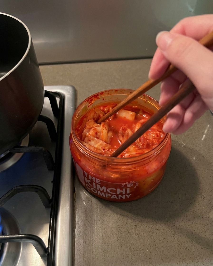 Essstäbchen heben eine Portion rotes, scharfes Kimchi aus einem klaren Glas.